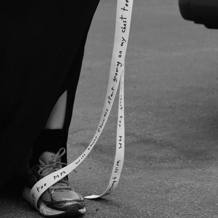 Black and white photo, wearing dad sneakers and a dress with slit. Mark Milroy canvas belt in natural colour with writing. cruba